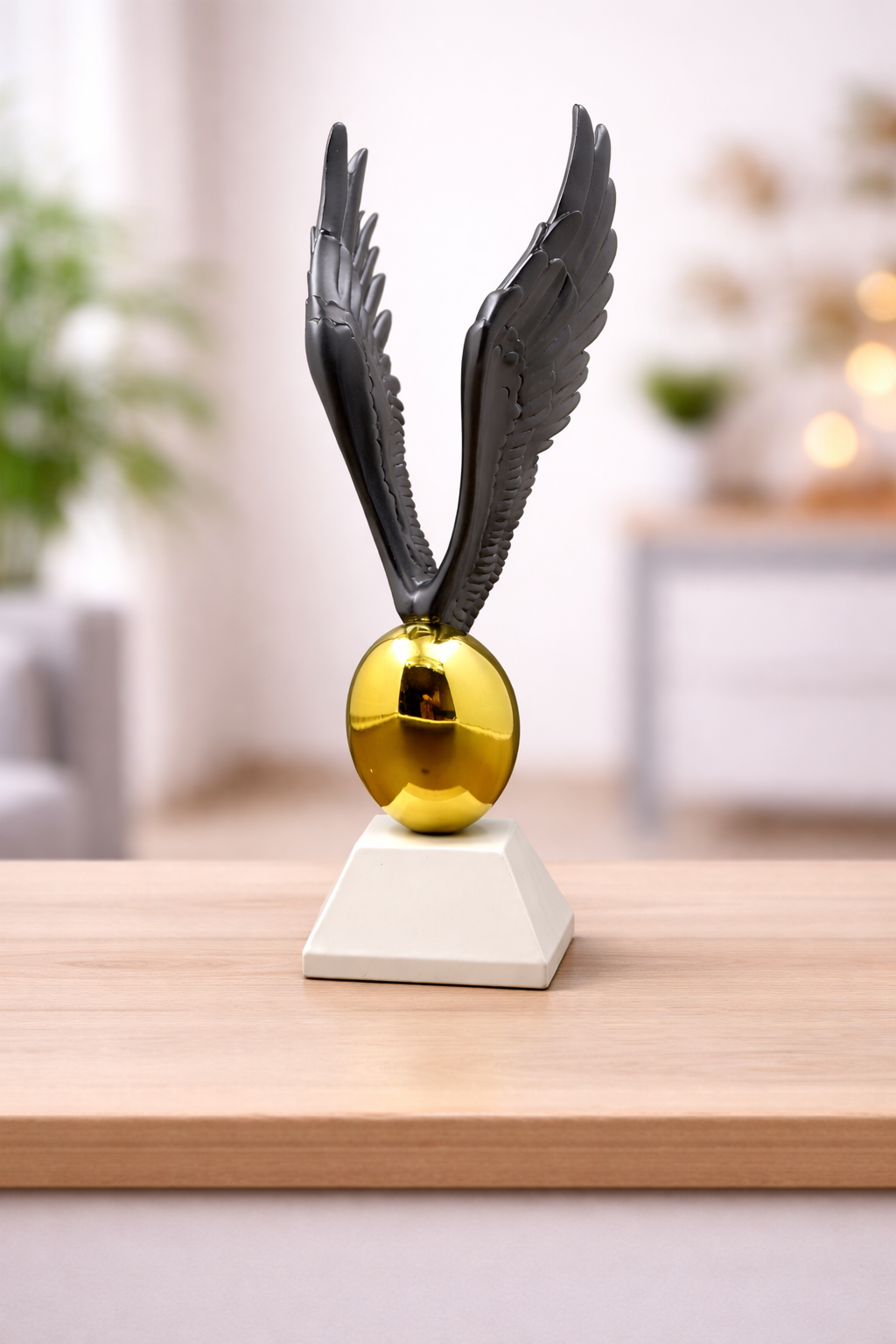Decorative object with black wings and gold sphere on a white base, placed on a wooden surface.