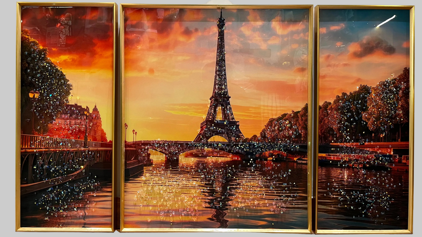 A three-panel framed wall art set depicting the Eiffel Tower at sunset over a river, with warm orange and red sky tones, dark tree silhouettes, and reflective water. The panels are enclosed in gold frames and shown hanging on a light wall.