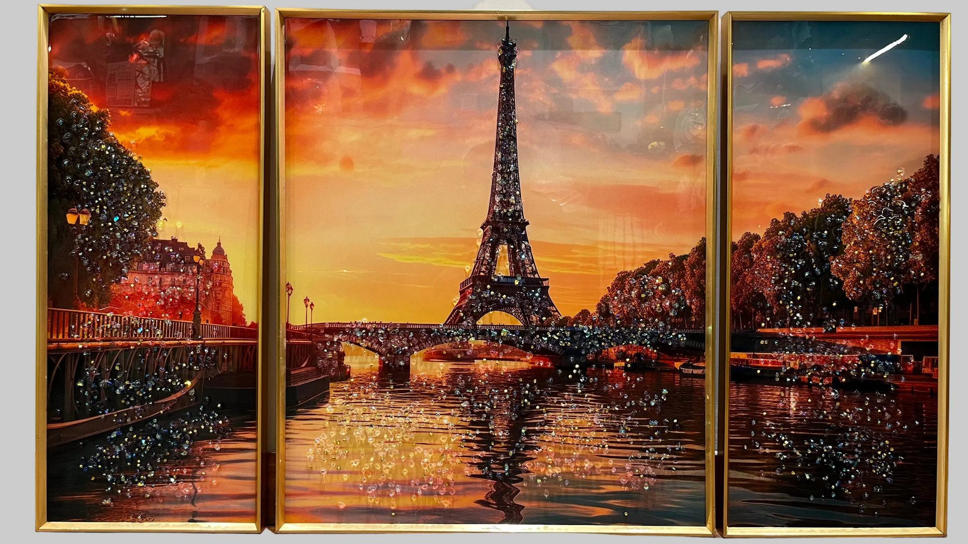 A three-panel framed wall art set depicting the Eiffel Tower at sunset over a river, with warm orange and red sky tones, dark tree silhouettes, and reflective water. The panels are enclosed in gold frames and shown hanging on a light wall.