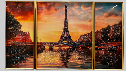 A three-panel framed wall art set depicting the Eiffel Tower at sunset over a river, with warm orange and red sky tones, dark tree silhouettes, and reflective water. The panels are enclosed in gold frames and shown hanging on a light wall.